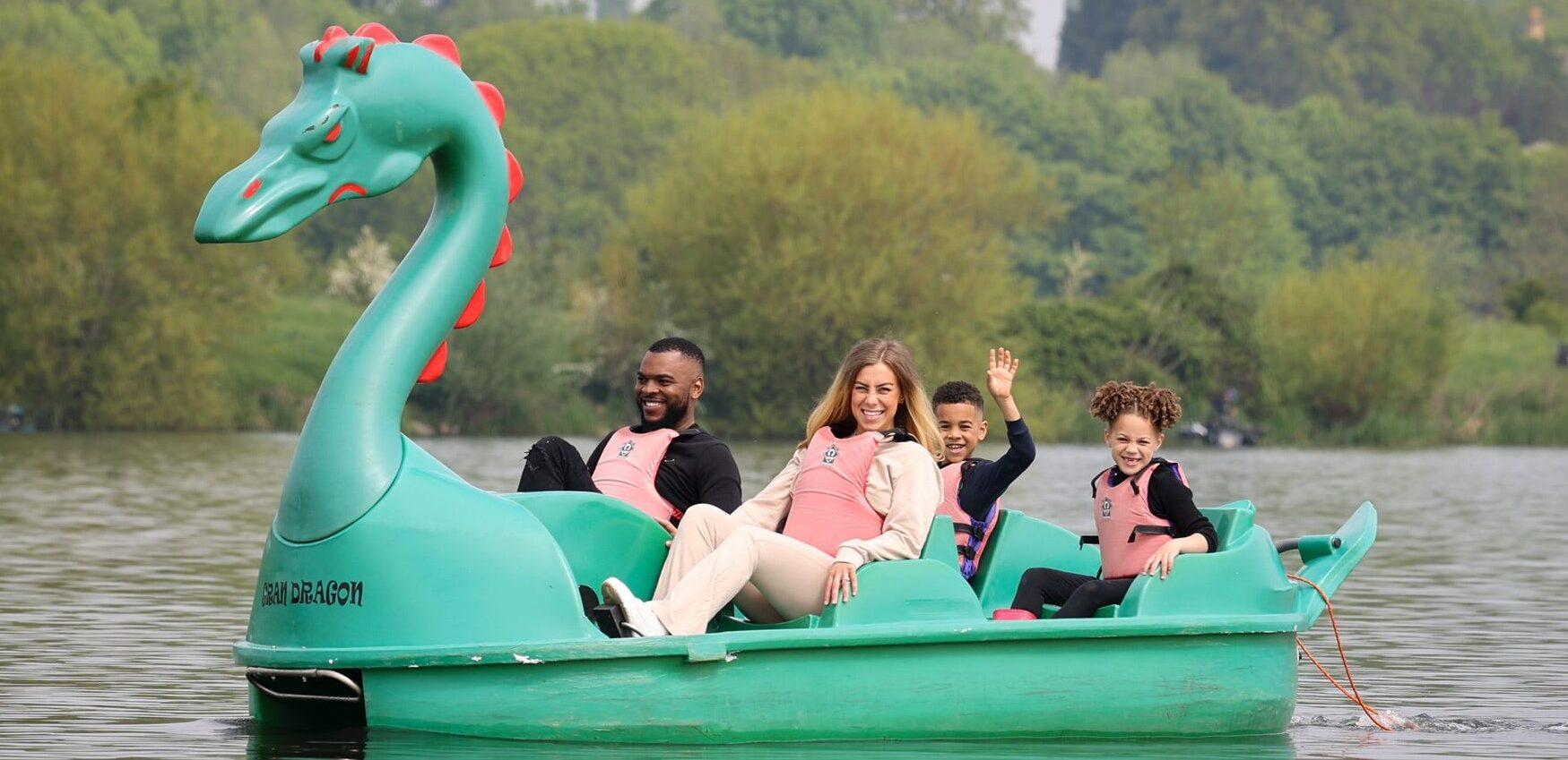 A group of two adults and two children are paddling in a green dragon pedalo on a lake with trees in the background