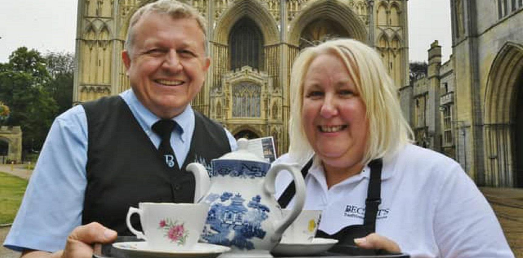 A couple standing in front of Peterborough Cathedral holding a tea tray